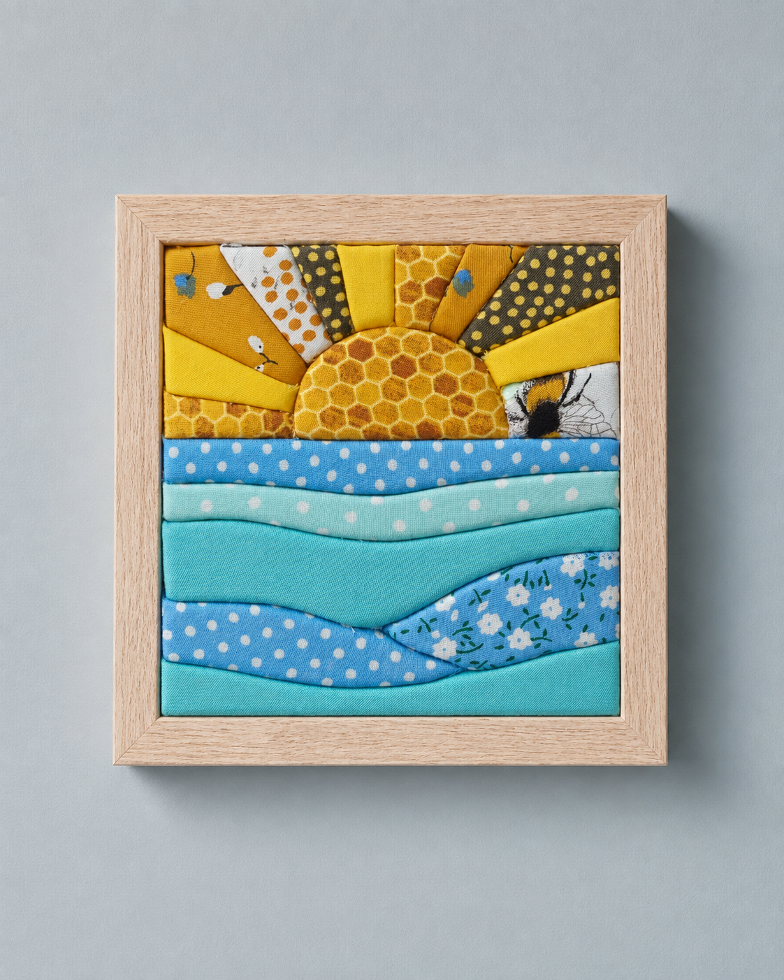 A square framed kinusaiga wall art piece depicting a sun over blue waves, with bees and honeycomb patterns. The artwork is housed in a light natural wood frame and photographed against a solid gray background.