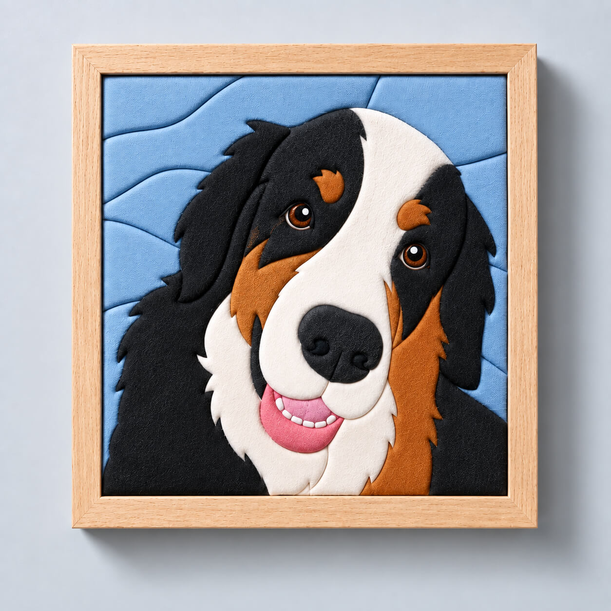 A custom kinusaiga pet portrait in a natural wood shadowbox. A Bernese mountain dog's face is built entirely from tucked fabric in black, cream and rust, against a pale blue background of overlapping fabric bands. The frame hangs on a soft gray wall.