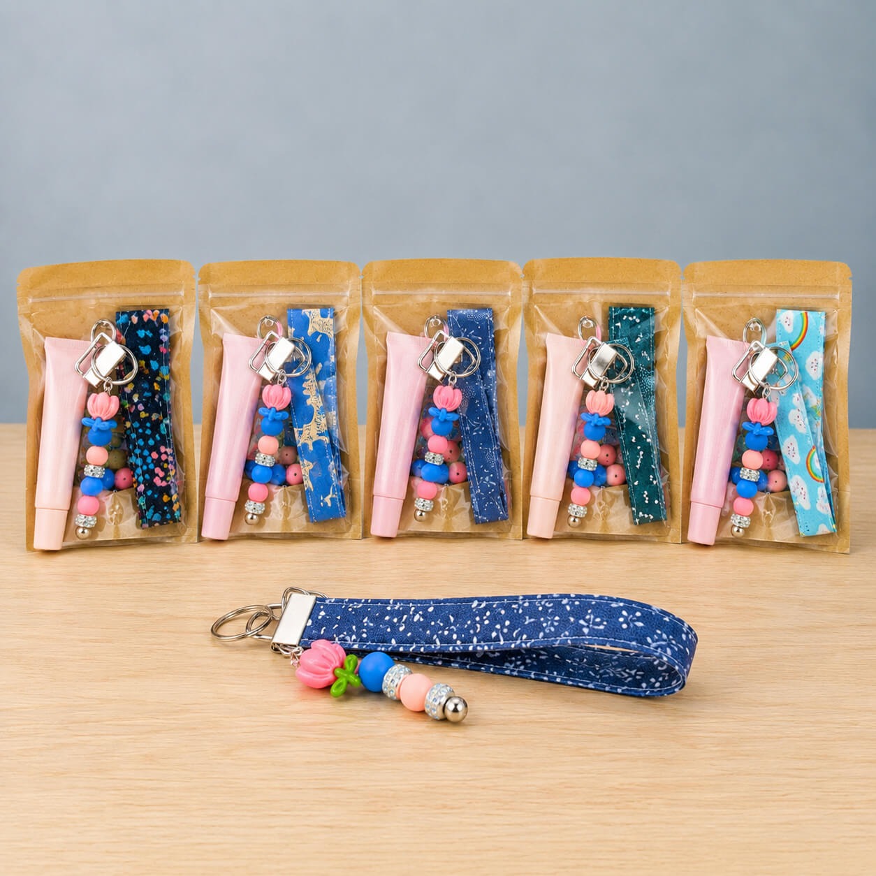 A row of bagged lip balm wristlet kits in cool prints, each holding a pink lip balm tube, a beaded charm and a folded fabric strap, with a finished navy floral wristlet laid out in front