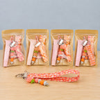 A row of bagged lip balm wristlet kits in warm prints, each holding a pink lip balm tube, a beaded charm and a folded fabric strap, with a finished orange and pink wristlet laid out in front