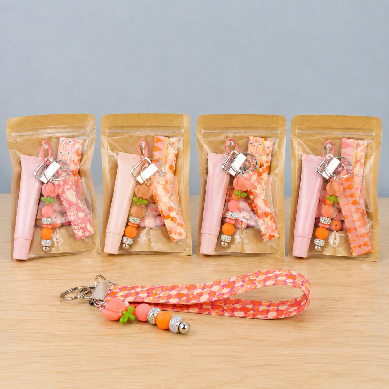A row of bagged lip balm wristlet kits in warm prints, each holding a pink lip balm tube, a beaded charm and a folded fabric strap, with a finished orange and pink wristlet laid out in front