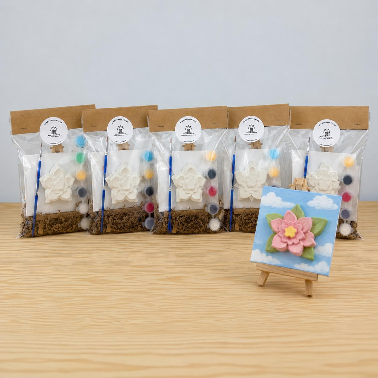 Five Make It Mine easel kits in the Flower design, each bagged with a kraft header card, with a finished painted flower on a mini canvas standing on its easel in front