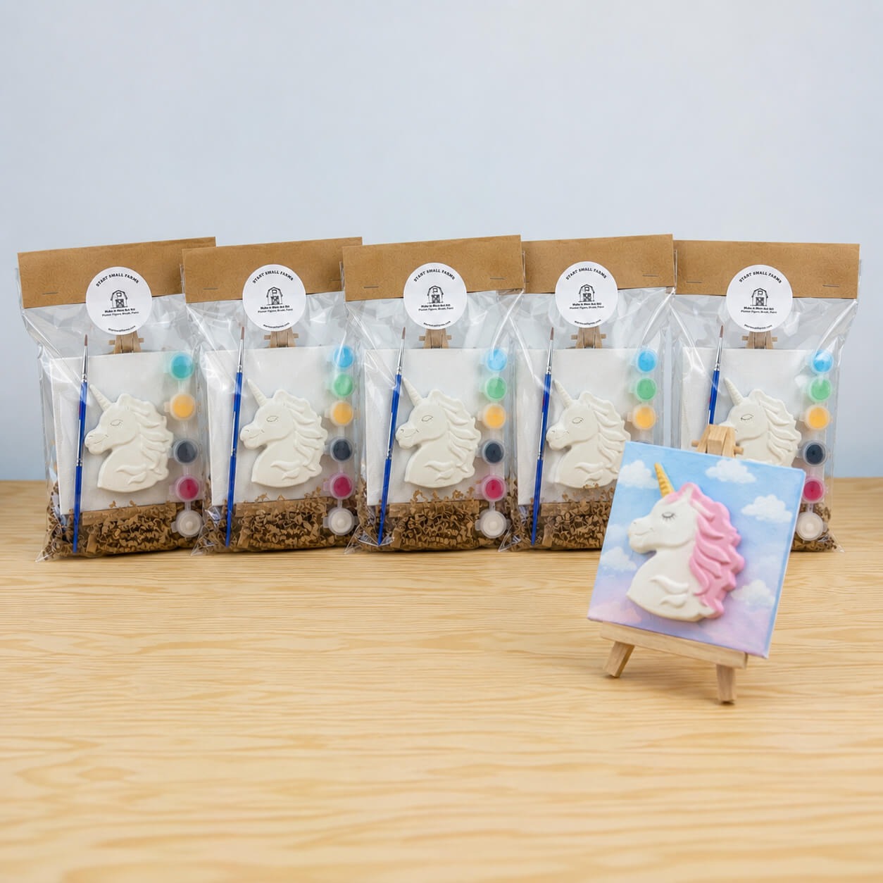 Five Make It Mine easel kits in the Unicorn design, each bagged with a kraft header card, with a finished painted unicorn on a mini canvas standing on its easel in front