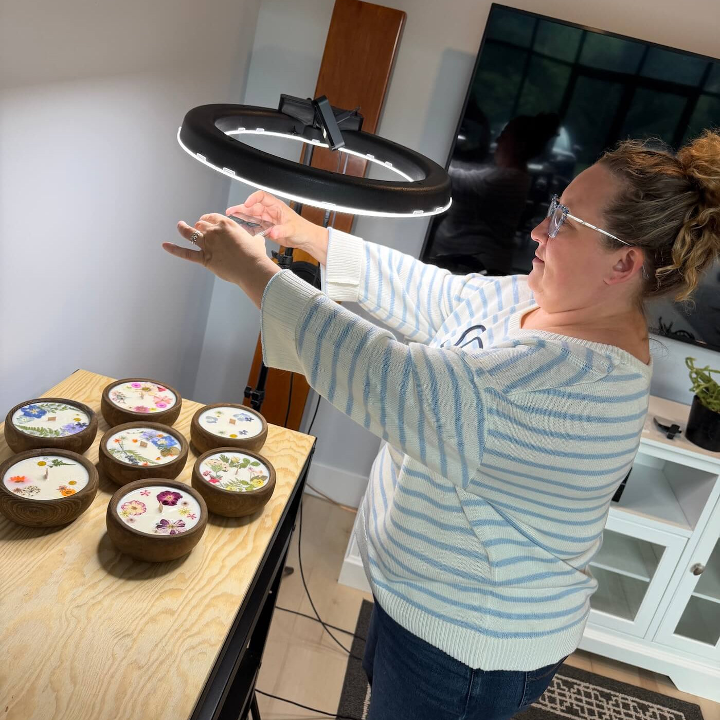 A woman in a light blue and white striped sweater adjusts a black ring light above six small round wooden bowls containing colorful wax melts or soaps, set in a home office environment.