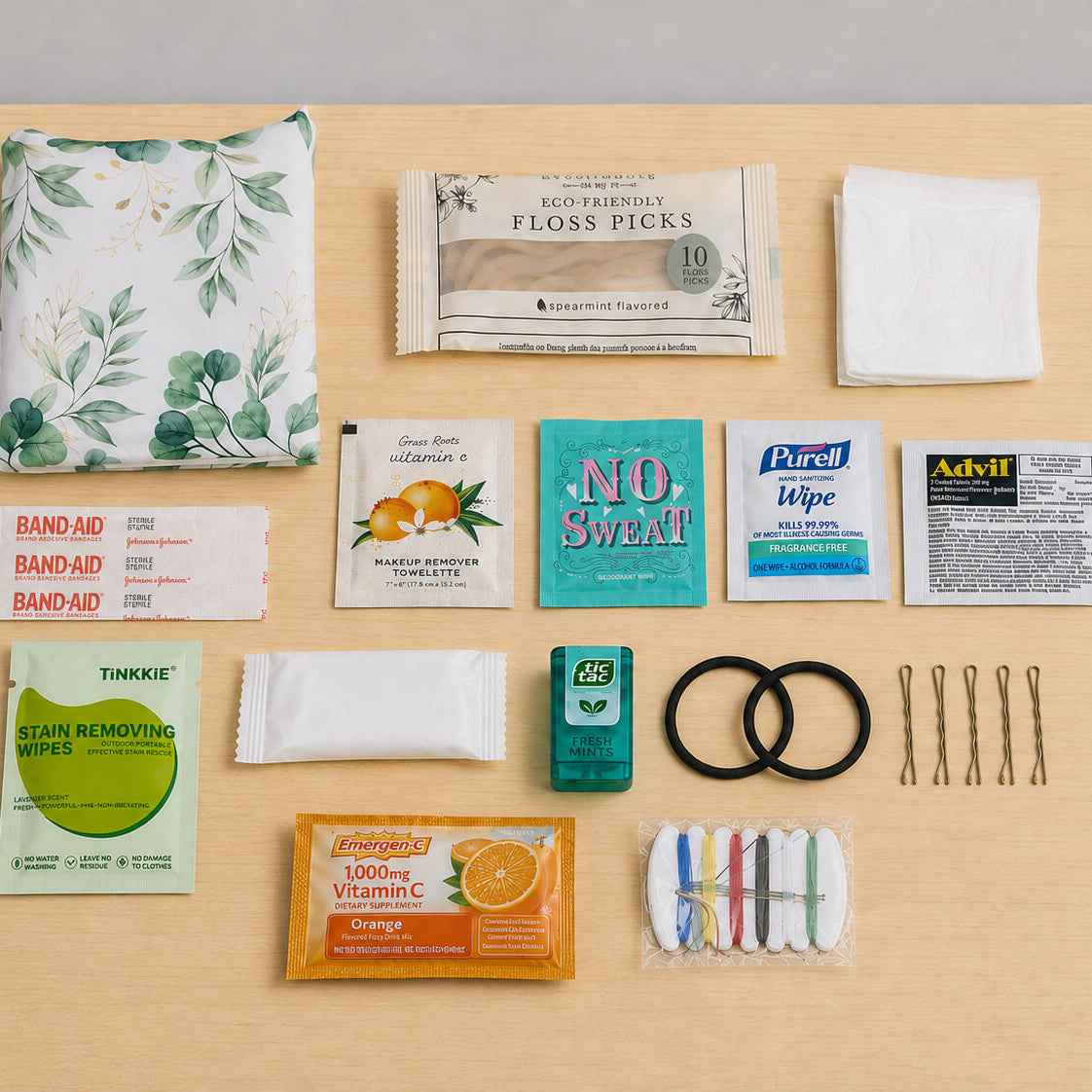 A flat-lay arrangement of hair and beauty travel accessories on a light wood surface, including a green-and-white leaf-patterned pouch, floss picks, band-aids, wipes, a small teal case, bobby pins, and other compact-size toiletries.