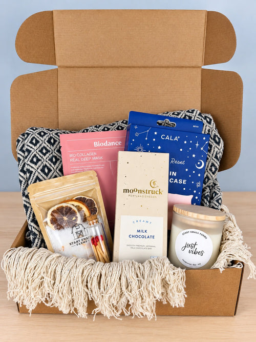 The Retreat in Place box open in kraft, lined with a navy and cream diamond-weave cotton throw: a Start Small Farms bath soak pouch with dried citrus slices, a corked vial of wooden matches, a Biodance collagen sheet mask, a CALA satin pillowcase in its blue box, a Moonstruck milk chocolate bar and the Start Small Farms Just Vibes soy candle with a wooden lid.