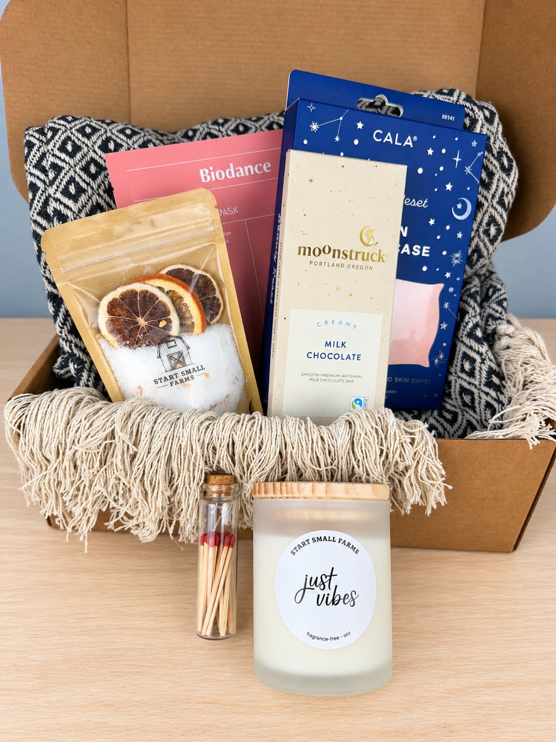 The Start Small Farms Just Vibes candle in a frosted glass jar and a corked vial of wooden matches in front of the open box.