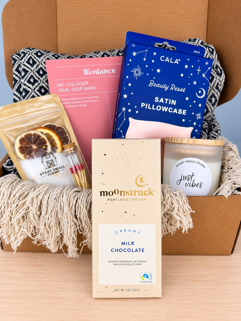 The Moonstruck creamy milk chocolate bar in its speckled cream wrapper, standing in front of the open box with the candle, matches and bath soak behind it.