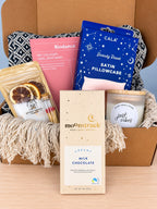 The Moonstruck creamy milk chocolate bar in its speckled cream wrapper, standing in front of the open box with the candle, matches and bath soak behind it.