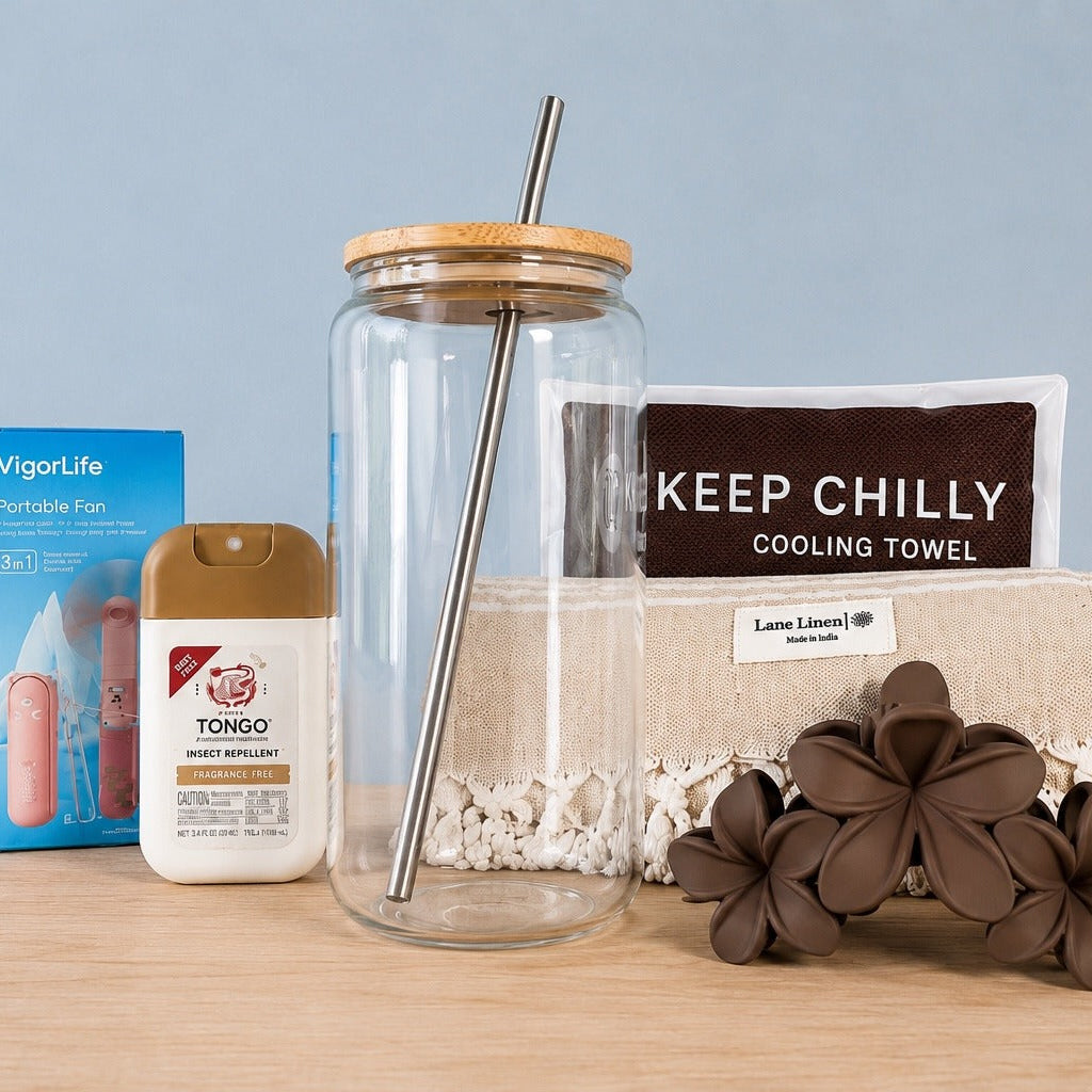 The Hot Girl Kit contents arranged on a wood surface against a pale blue backdrop: a portable fan in its box, a Tongo fragrance-free insect repellent pump, a 16oz glass tumbler with bamboo lid and metal straw, a Keep Chilly cooling towel in its pouch, a fringed Turkish towel and a taupe plumeria claw clip
