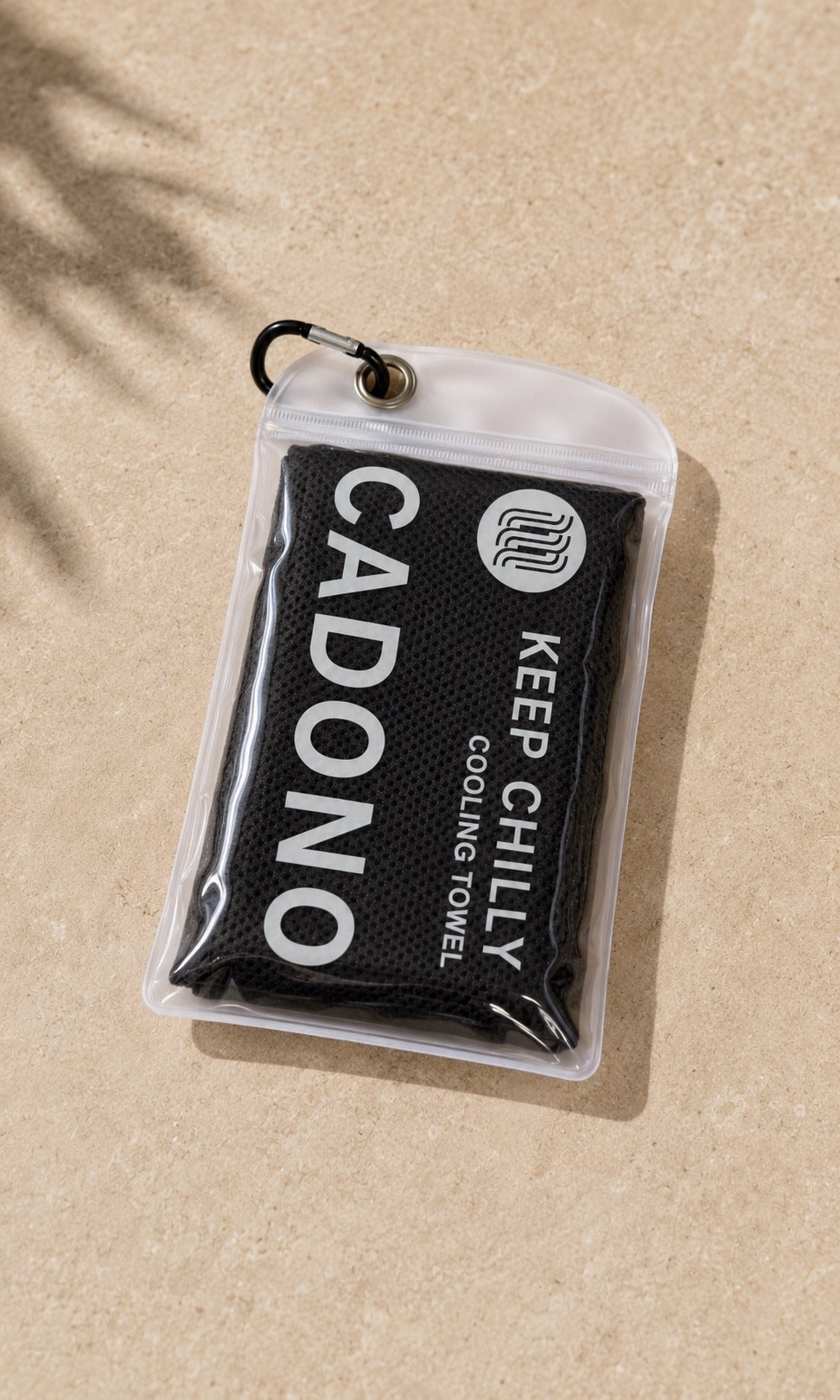 The Keep Chilly cooling towel folded inside its clear zip pouch with a carabiner clip, on sand under palm shadow