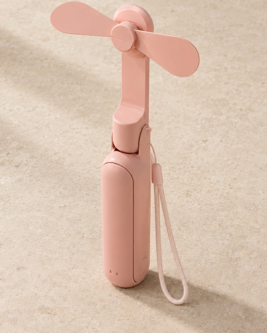 The portable fan in blush pink, unfolded and standing upright with its wrist lanyard, on pale stone