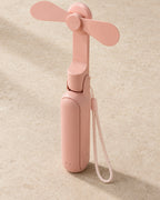 The portable fan in blush pink, unfolded and standing upright with its wrist lanyard, on pale stone