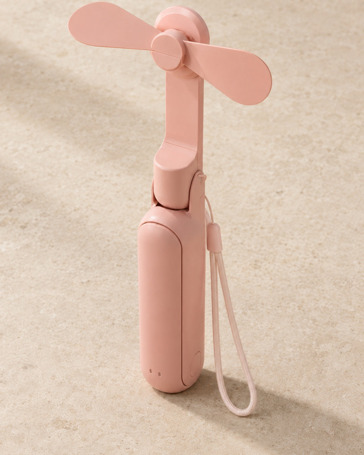 The portable fan in blush pink, unfolded and standing upright with its wrist lanyard, on pale stone