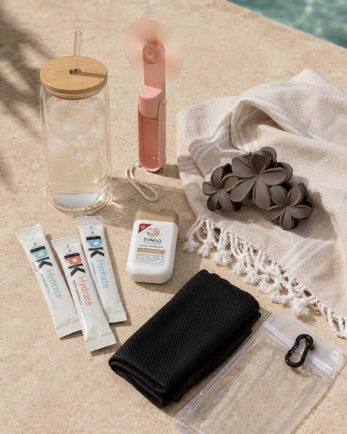The Hot Girl Kit laid out on stone at the edge of a pool: the glass tumbler filled with ice, the blush pink folding fan, three Pure Kick hydration sticks, Tongo insect repellent, a folded black cooling towel with its clear pouch, a fringed Turkish towel and a taupe plumeria claw clip