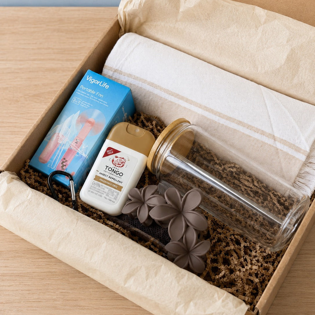 The Hot Girl Kit packed in a kraft gift box with crinkle paper: a VigorLife portable fan in its blue box, a Tongo insect repellent pump, a glass tumbler with bamboo lid, brown plumeria hair clips and a fringed Turkish towel