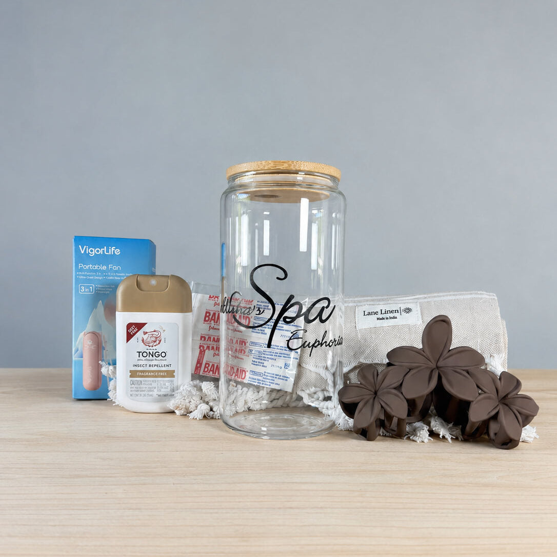 The Hot Girl Kit contents lined up on a wood surface against a pale blue backdrop: a VigorLife portable fan box, Tongo insect repellent, a glass tumbler with bamboo lid and metal straw, a Keep Chilly cooling towel, wildflower bandages, a fringed Turkish towel and brown plumeria hair clips
