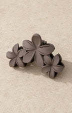 A taupe plumeria flower claw hair clip on pale stone, three matte flowers set across the clip