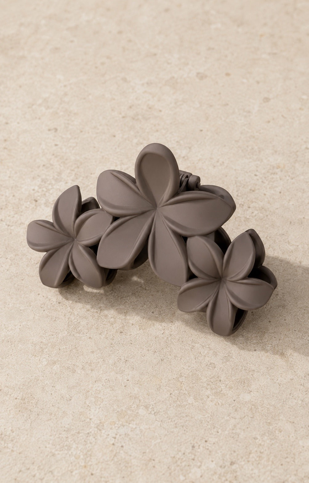 A taupe plumeria flower claw hair clip on pale stone, three matte flowers set across the clip