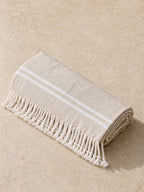The Turkish beach towel rolled up on pale stone, sand colored with white stripes and a hand-knotted fringe
