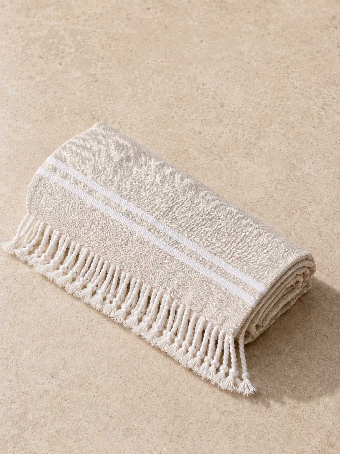 The Turkish beach towel rolled up on pale stone, sand colored with white stripes and a hand-knotted fringe