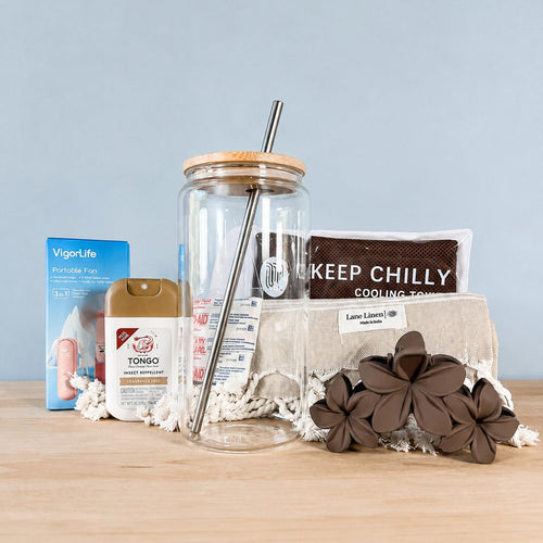 The Hot Girl Kit contents lined up on a wood surface against a pale blue backdrop: a VigorLife portable fan box, Tongo insect repellent, a glass tumbler with bamboo lid and metal straw, a Keep Chilly cooling towel, wildflower bandages, a fringed Turkish towel and brown plumeria hair clips