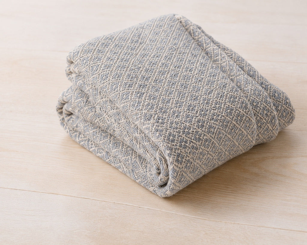 The cotton throw blanket folded on pale wood, woven in a blue and cream diamond pattern