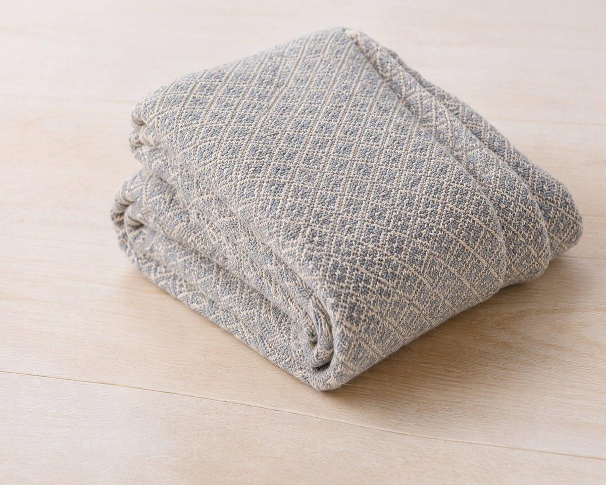 The cotton throw blanket folded on pale wood, woven in a blue and cream diamond pattern