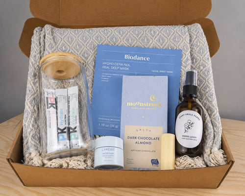 The Treat Yourself box packed in kraft with the cotton throw as a nest: a 16oz glass tumbler with bamboo lid holding three Pure Kick hydration sticks, a Biodance collagen sheet mask, a Moonstruck chocolate bar, a Laneige Water Bank lip sleeping mask, an EOS lip balm and the Start Small Farms Fresh Linen room and linen spray