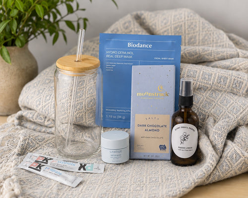 The Treat Yourself contents styled on the cotton throw beside a potted plant: the glass tumbler with bamboo lid and glass straw, a Biodance collagen sheet mask, a Moonstruck chocolate bar, a Laneige Water Bank lip sleeping mask, three Pure Kick hydration sticks and the Start Small Farms Fresh Linen room spray
