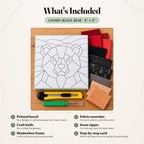 What's included in the Chunky Black Bear kit, laid out on a wood table: the printed bear board, fabric swatches in reds, blacks, greys and tan, a craft knife and a seam ripper. The shadowbox frame and the step-by-step card are not pictured.