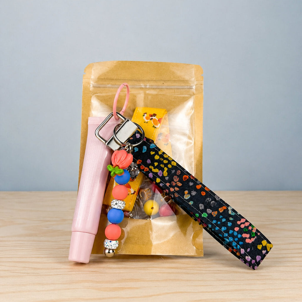 A gift set in a clear gold pouch on a light wood table, featuring a black wristlet strap with colorful heart patterns, a pink cylindrical item, and a beaded charm keychain.