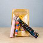 A gift set in a clear gold pouch on a light wood table, featuring a black wristlet strap with colorful heart patterns, a pink cylindrical item, and a beaded charm keychain.