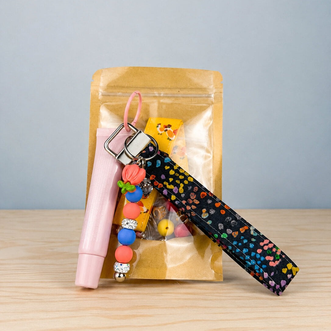 One lip balm wristlet kit in its kraft bag, with the pink lip balm tube, the beaded keychain bar and a black confetti print strap leaning against the front