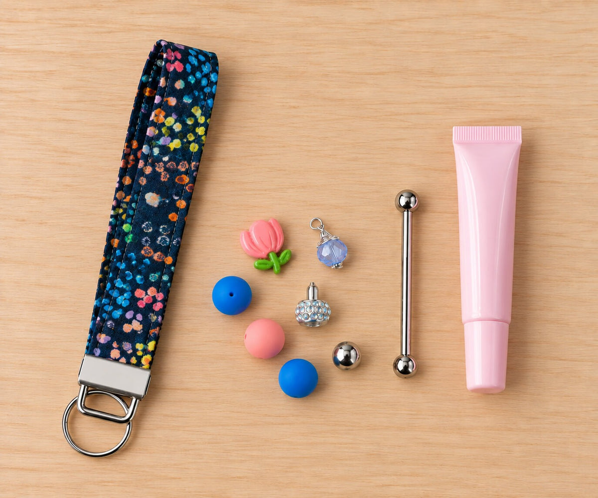 Everything in one lip balm wristlet kit laid flat on a light wood table: a navy wristlet strap in a colorful confetti floral print with a silver clasp and split ring, a pink lip balm tube, a silver beadable keychain bar, and a handful of beads and charms including a pink tulip, a rhinestone bead, and blue and pink silicone beads.