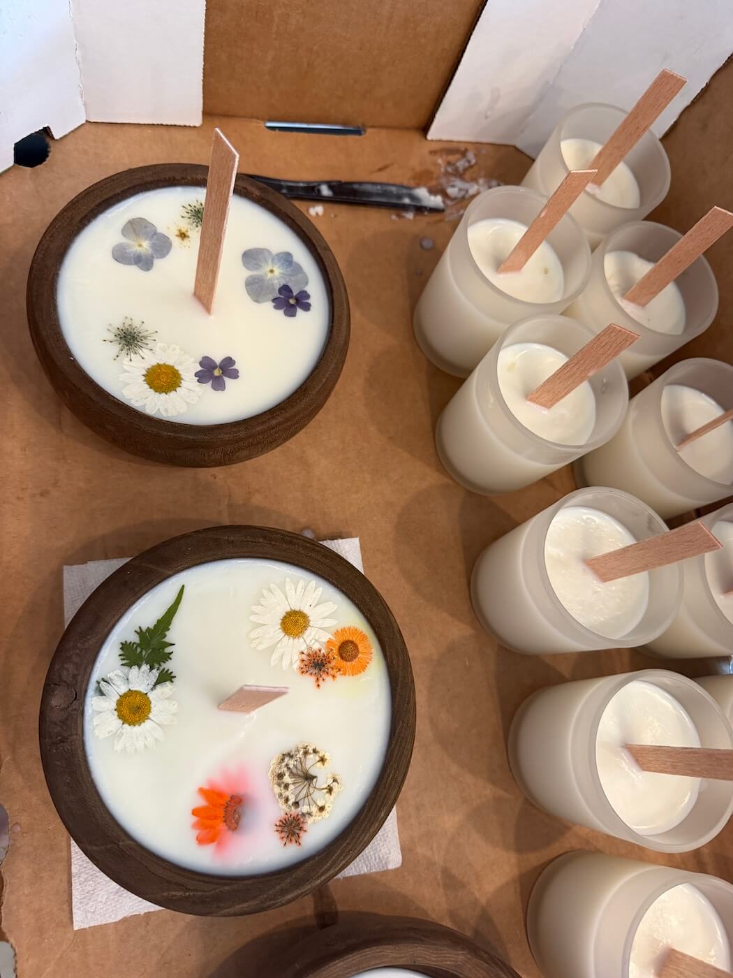 Overhead view of freshly poured soy candles drying in a box: two wooden bowl candles topped with pressed daisies, calendula, and ferns, beside a cluster of white glass jar candles with flat wooden wicks.