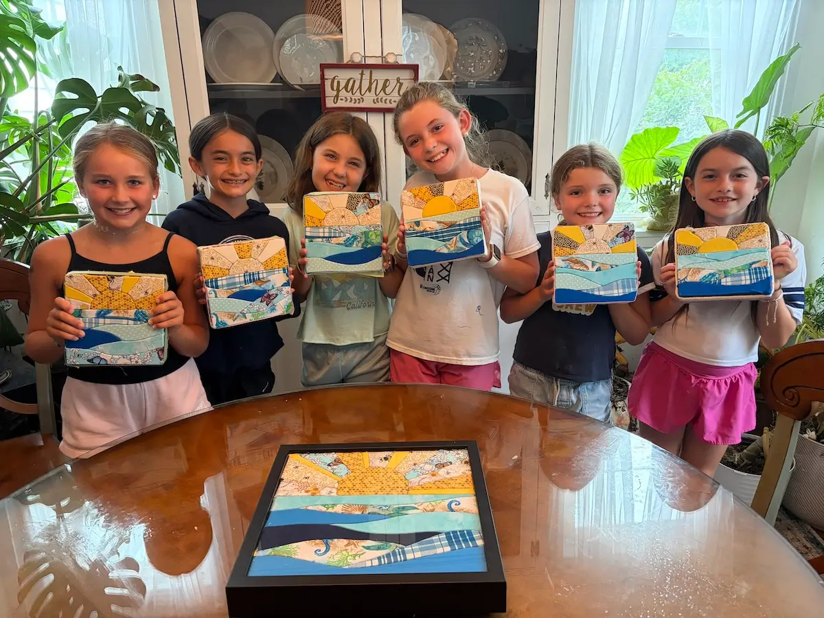 Six young makers standing side by side, each holding up a finished kinusaiga panel of a sun over water. Every panel follows the same design, but the fabric choices are different in each one, so the suns and the waves all read differently. A seventh framed piece sits on the wooden table in front of them.