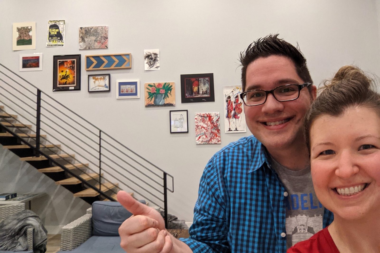 Kim and Jason smiling in a selfie while Jason points up at an eclectic gallery wall full of varied framed art beside a staircase.