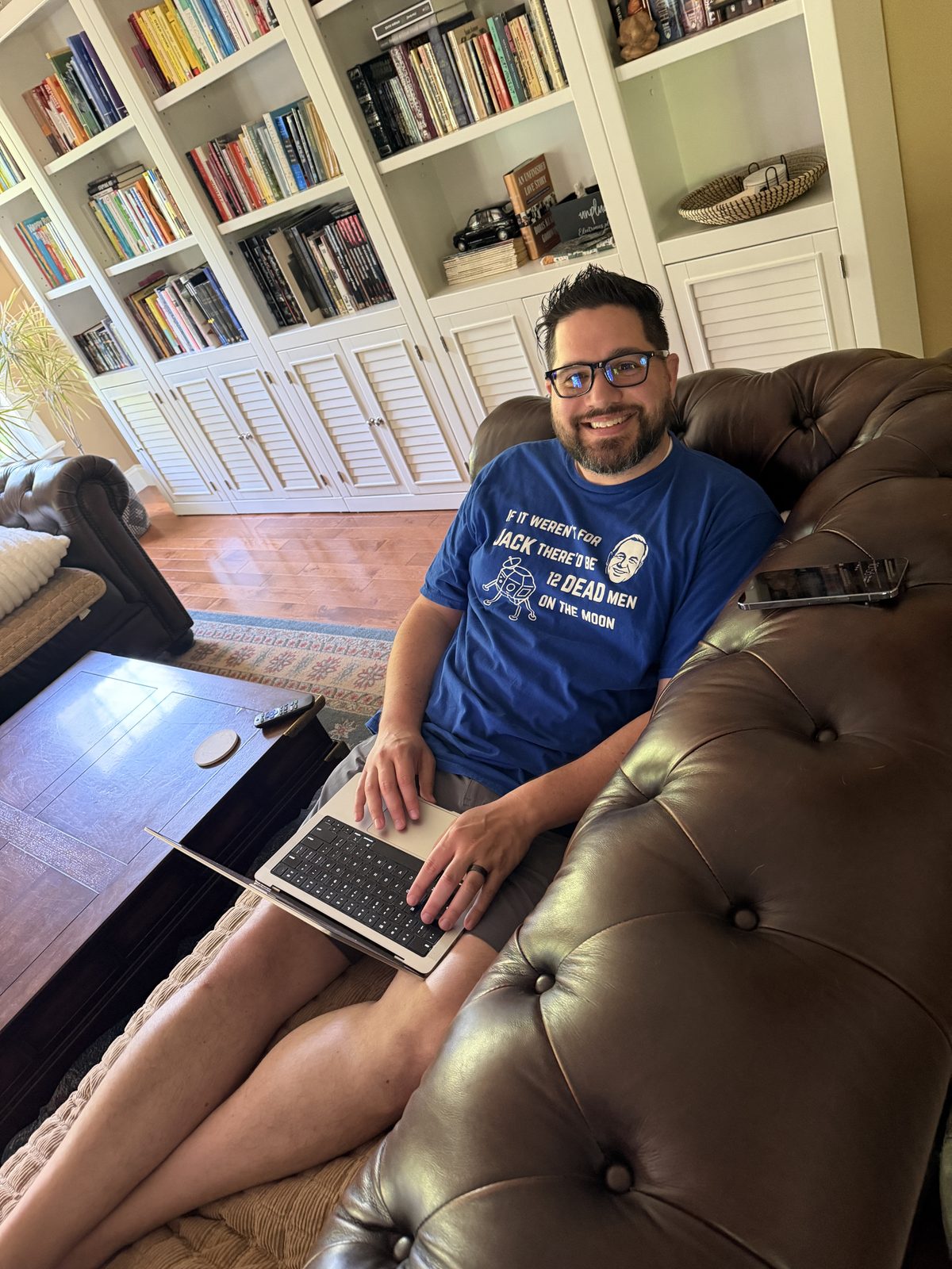 Jason sitting on a brown leather sofa with a laptop, smiling, in front of a full white bookshelf, wearing a blue t-shirt that reads 'If it weren't for Jack there'd be 12 dead men on the moon.'