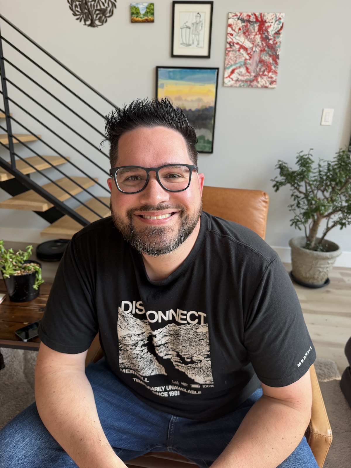 Jason sitting in a leather chair at the office, smiling in glasses and a black Merrell 'Disconnect' t-shirt, with the eclectic gallery wall and floating staircase behind him.