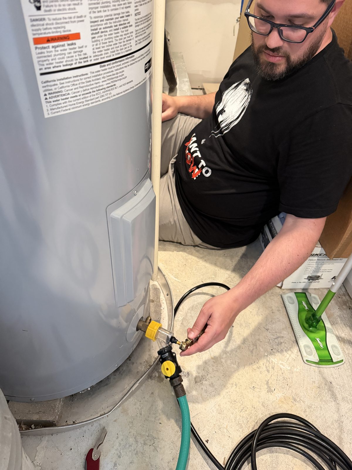 Jason kneeling beside a water heater, draining it through a clear inline sediment trap connected to a green garden hose.