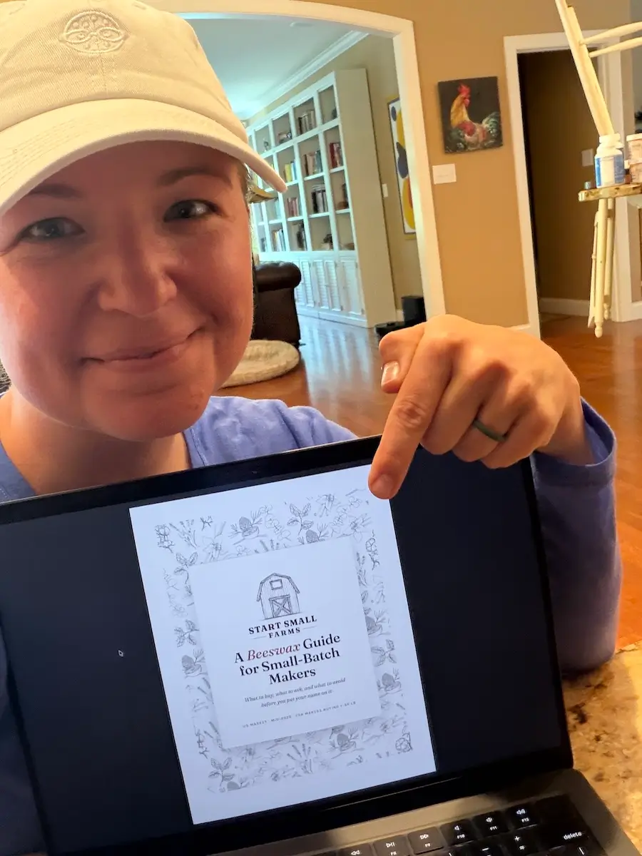 Kim in a cream ball cap, smiling and pointing at a laptop screen showing the cover of the Start Small Farms beeswax guide.