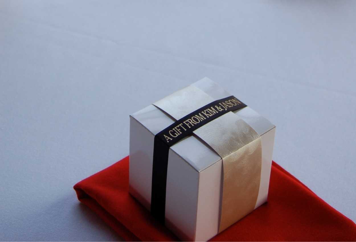 A photo of a wedding favor box with a gold strip of fabric and a black ribbon that says 'A Gift From Kim and Jason'.