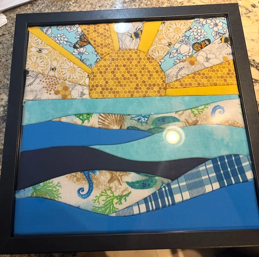 A finished kinusaiga piece in a black square frame: a sun rising over water, built entirely from fabric scraps tucked into grooves. The sun is golden honeycomb-print fabric, its rays alternating between solid yellow and prints of bees, butterflies, and white flowers on pale aqua. The water below is a stack of wavy bands in teal, cobalt, and navy, mixed with prints of seashells, seahorses, and green coral, plus one band of blue-and-white plaid.