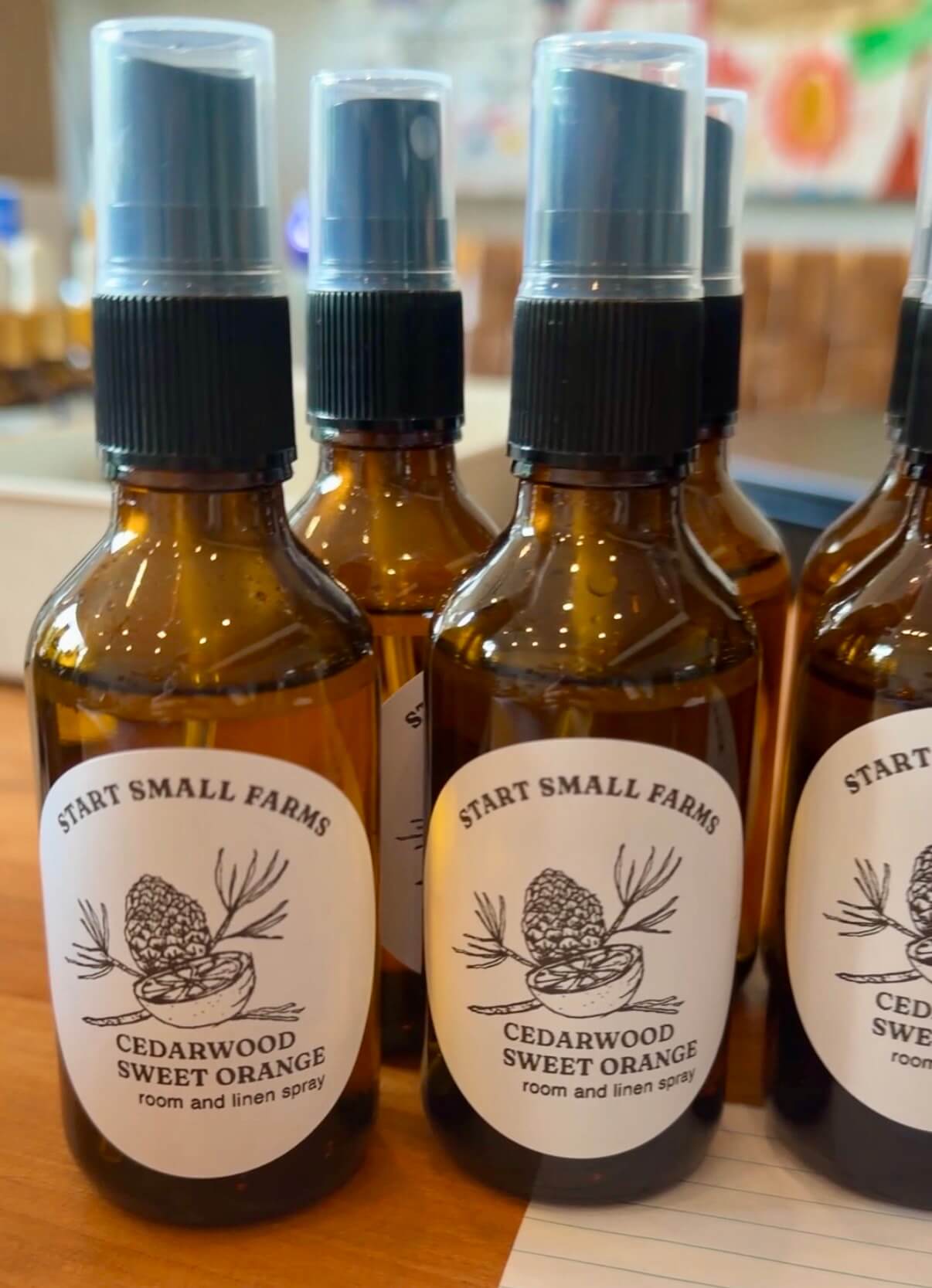 Several amber glass spray bottles with a Start Small Farms label reading 'Cedarwood Sweet Orange room and linen spray,' with a line-drawn logo of a pinecone, cedar sprig, and orange slice.